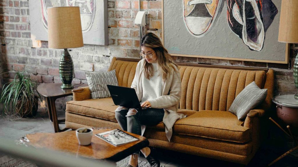 A young woman sits on a couch and works on her laptop